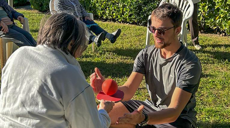 Le coach d'activité physique adapté de face guide une participante lors d'un exercice de coordination, pendant que les autres participants travaillent en autonomie.